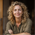 Smiling woman with curly blond hair in an olive shirt, leaning forward with arms crossed against a dark background