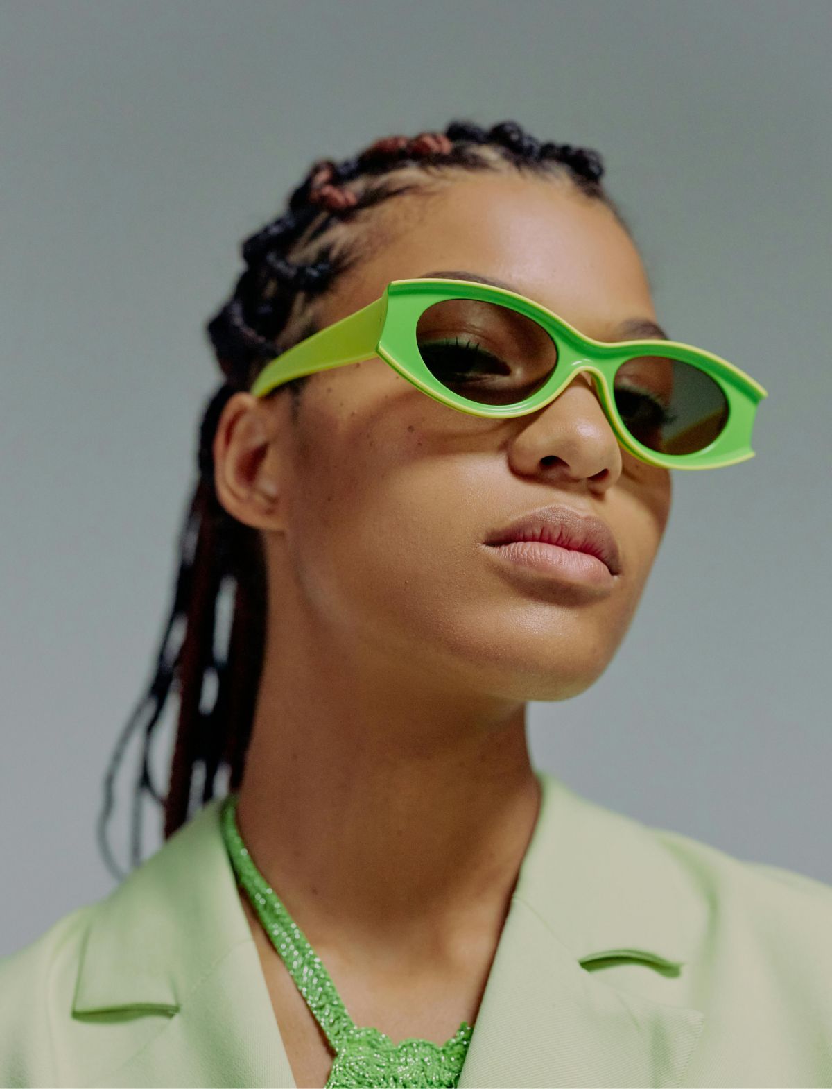 Woman with dark skin, wearing neon green sunglasses and blazer; braids.