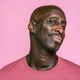 Man with shaved head, smiling and looking slightly to the side, wearing a pink shirt against a pink background.