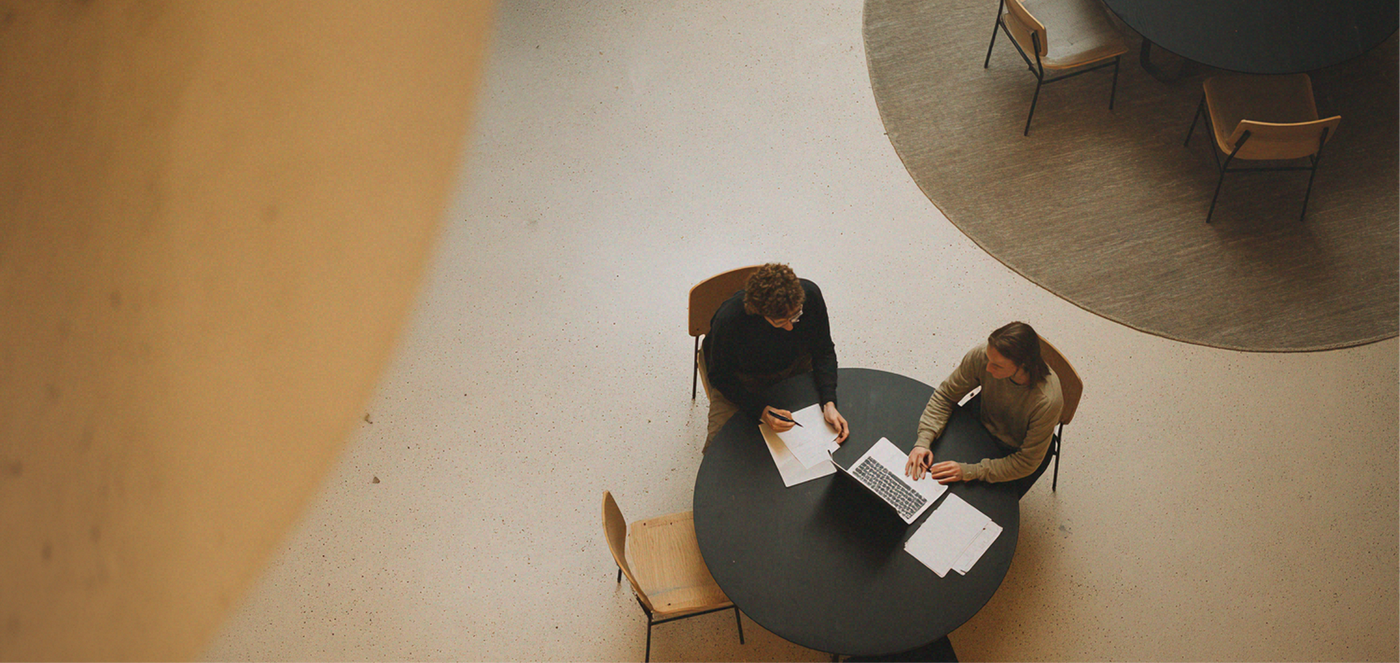 Twee mensen die documenten beoordelen aan een ronde tafel in een lichte lobby, gezien vanuit een bovenaanzicht.