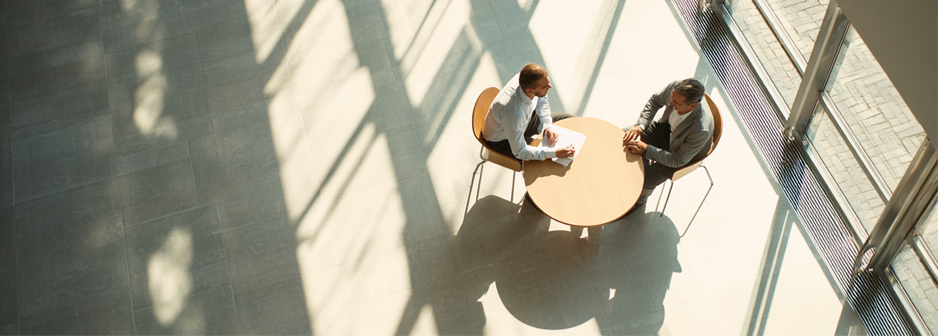 Twee mensen zitten aan een ronde tafel in een zonovergoten atrium, met schaduwen op de vloer.
