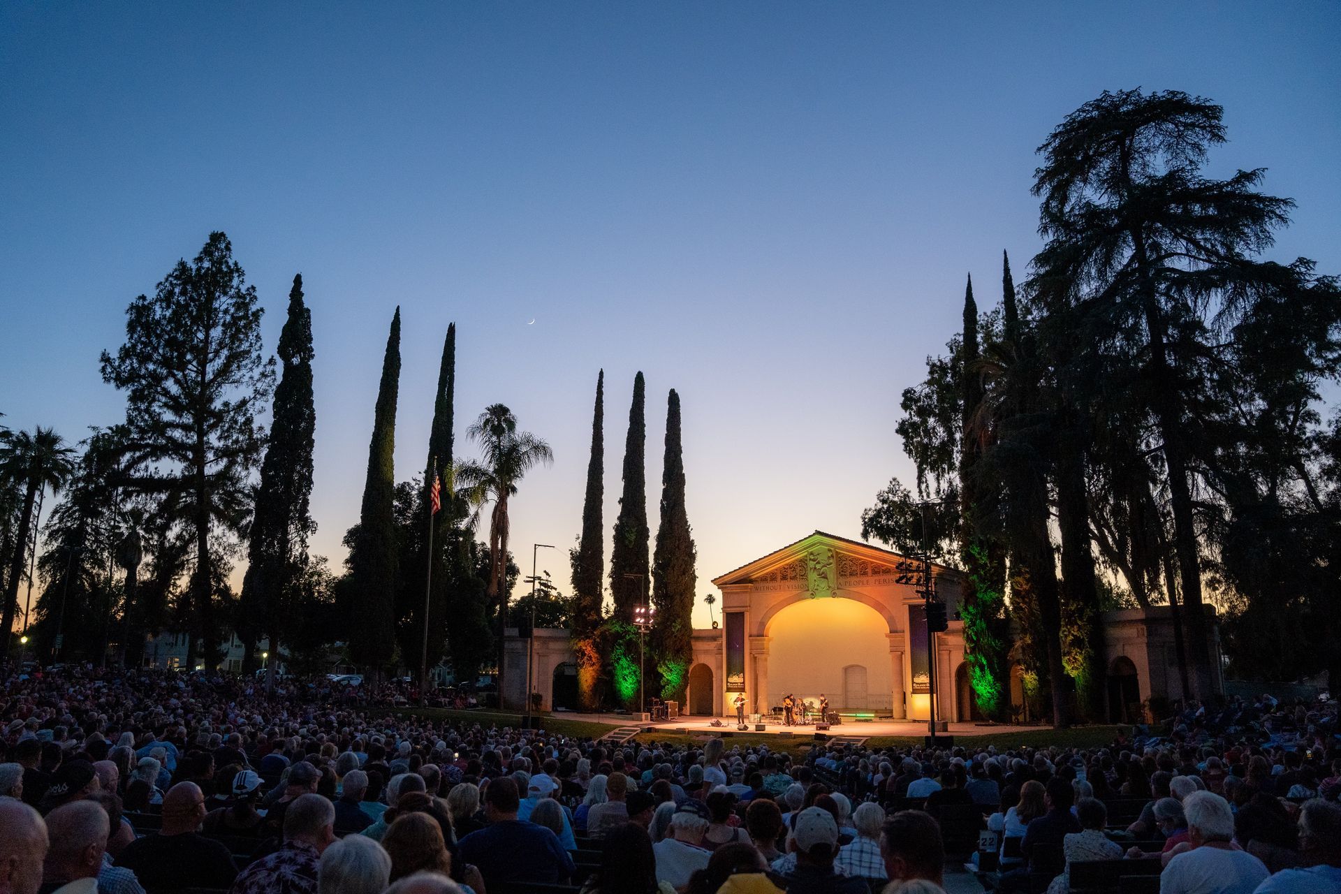 Outdoor concert at dusk: stage with performers, crowd seated on a lawn, tall trees, and architectural elements.