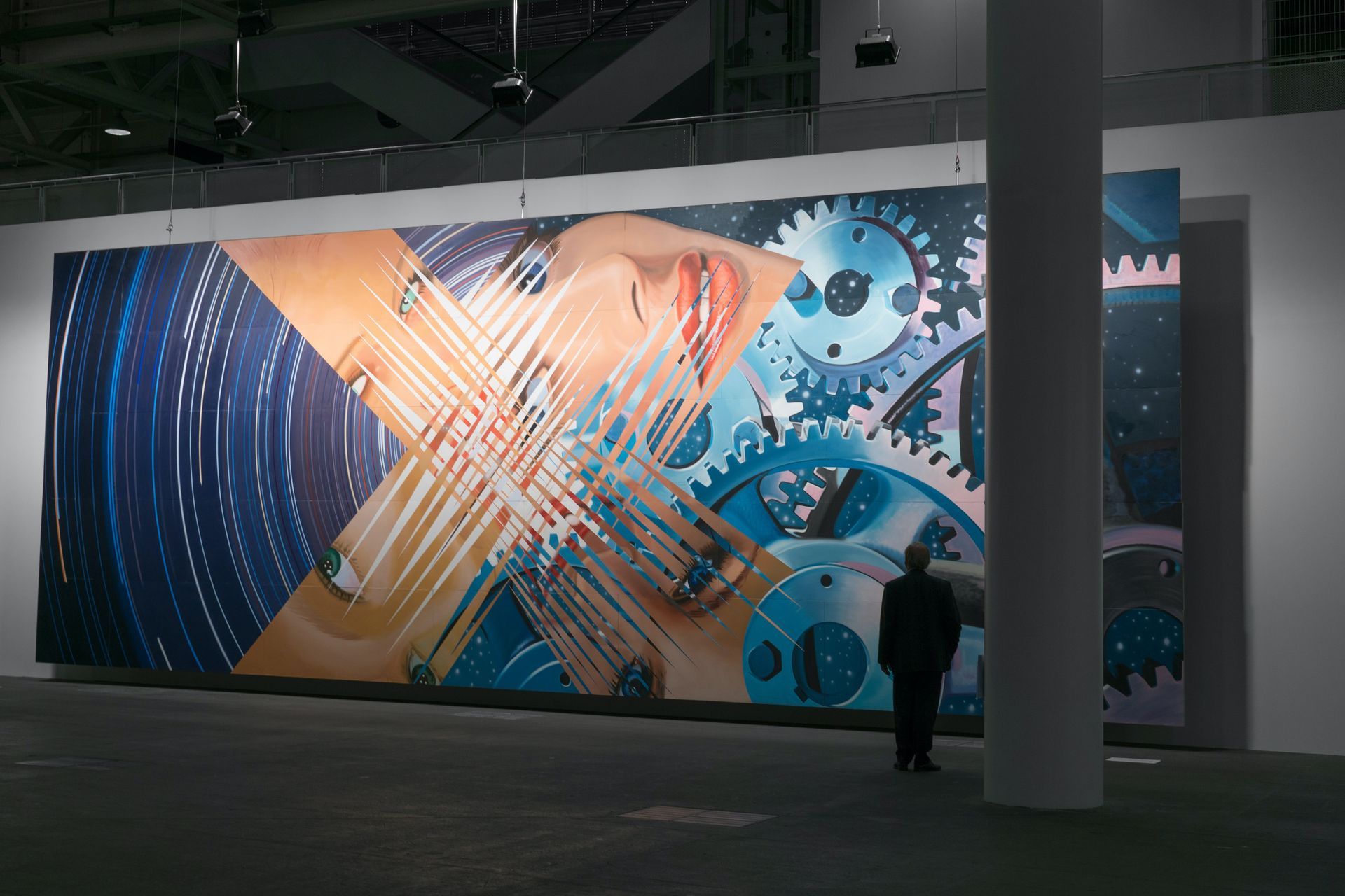 Large, abstract art display: colorful gears and face elements, man in silhouette, gallery setting.