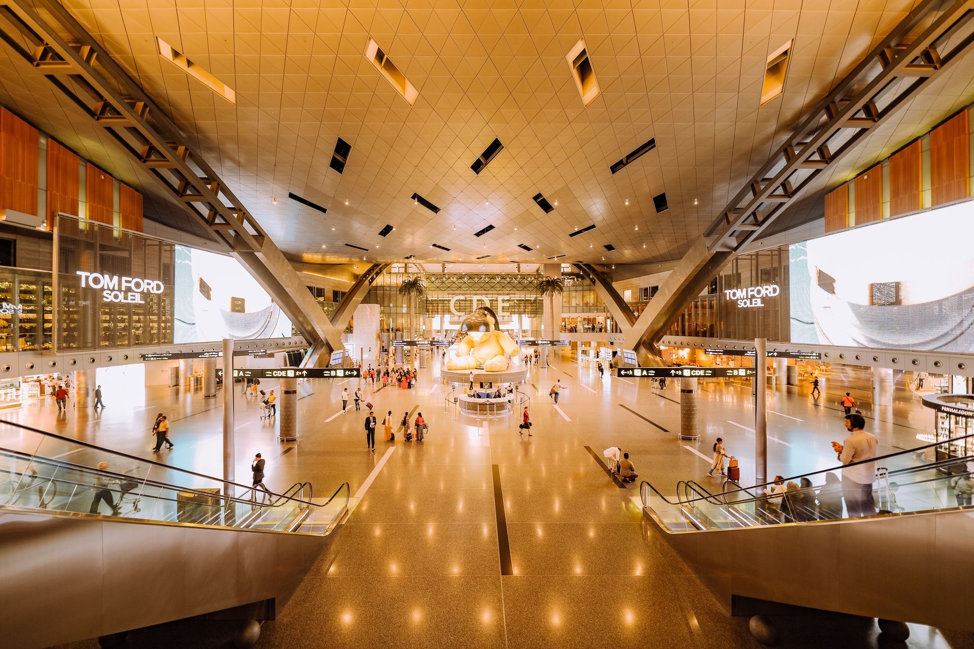 Spacious airport interior with modern design, people walking, escalators, and shops.