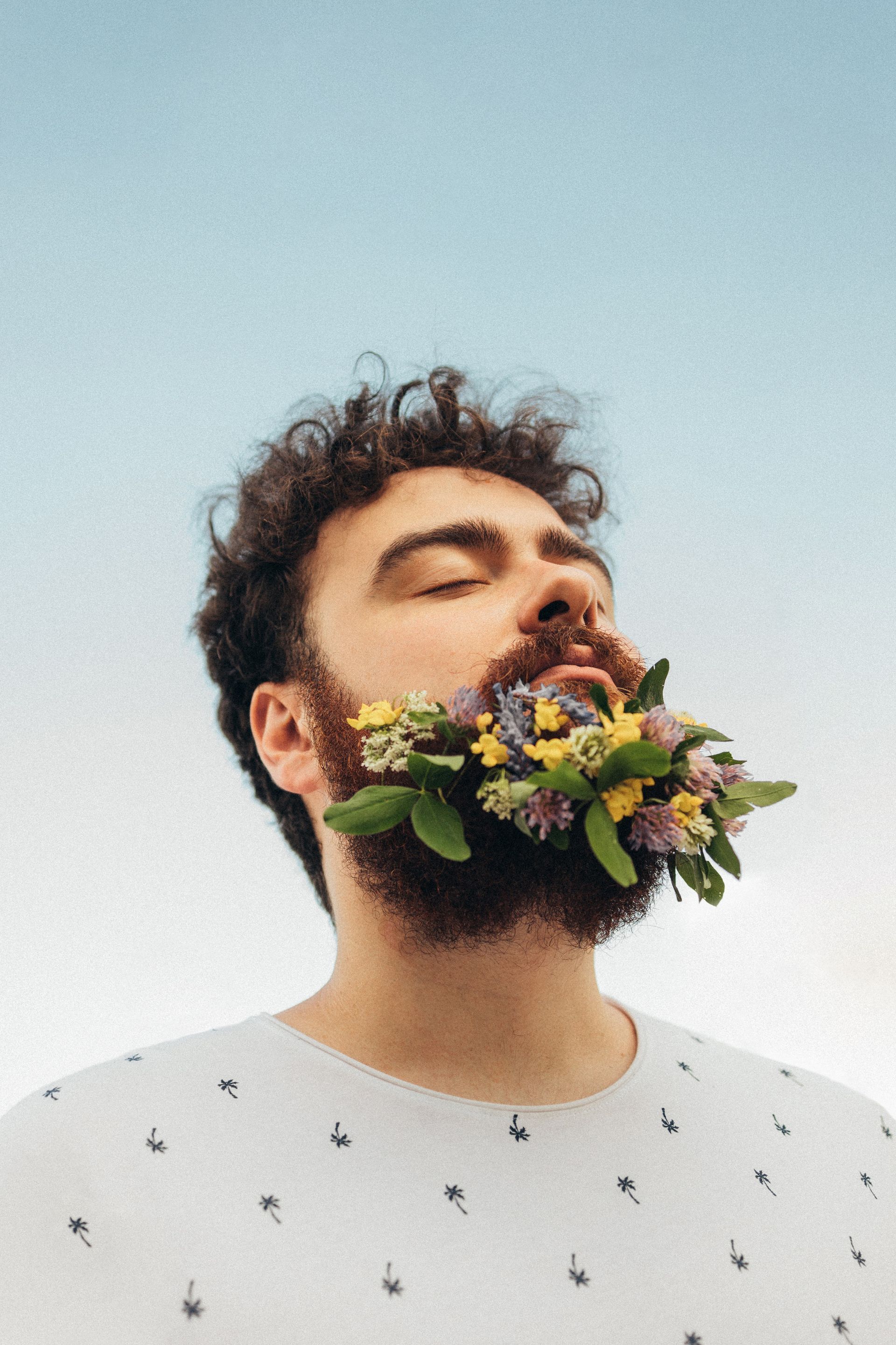 Man with curly hair and a beard filled with colorful flowers; against a blue sky.