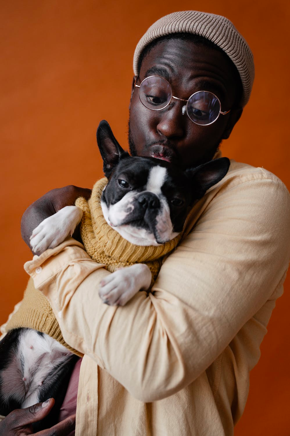 Man in tan shirt and hat holds and kisses a Boston Terrier dog in a yellow sweater, orange background.