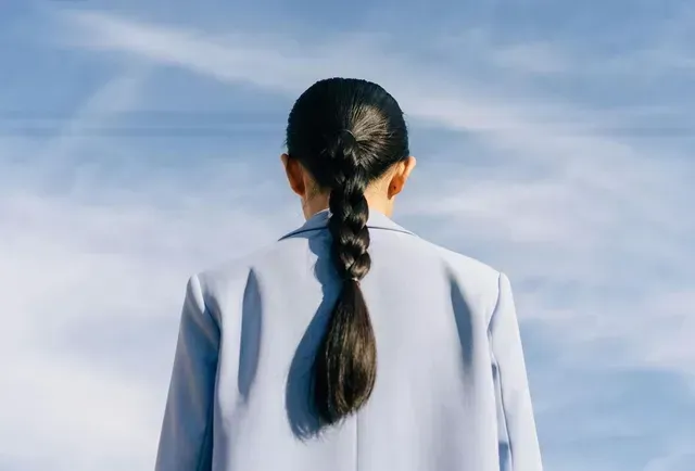 Woman in a light blue suit, back to the camera, with a long braid, against a blue sky.
