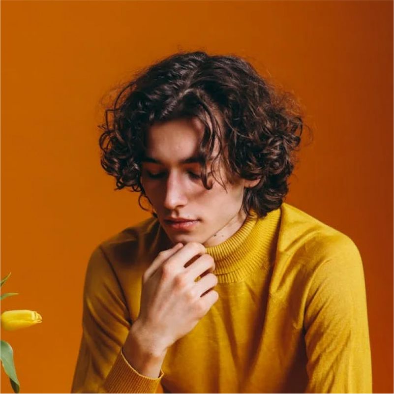 A person with curly brown hair wearing a yellow turtleneck, looking down with a hand on their chin. Orange background.