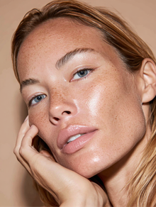 Woman with freckles and dewy skin, hand near face, looking up.