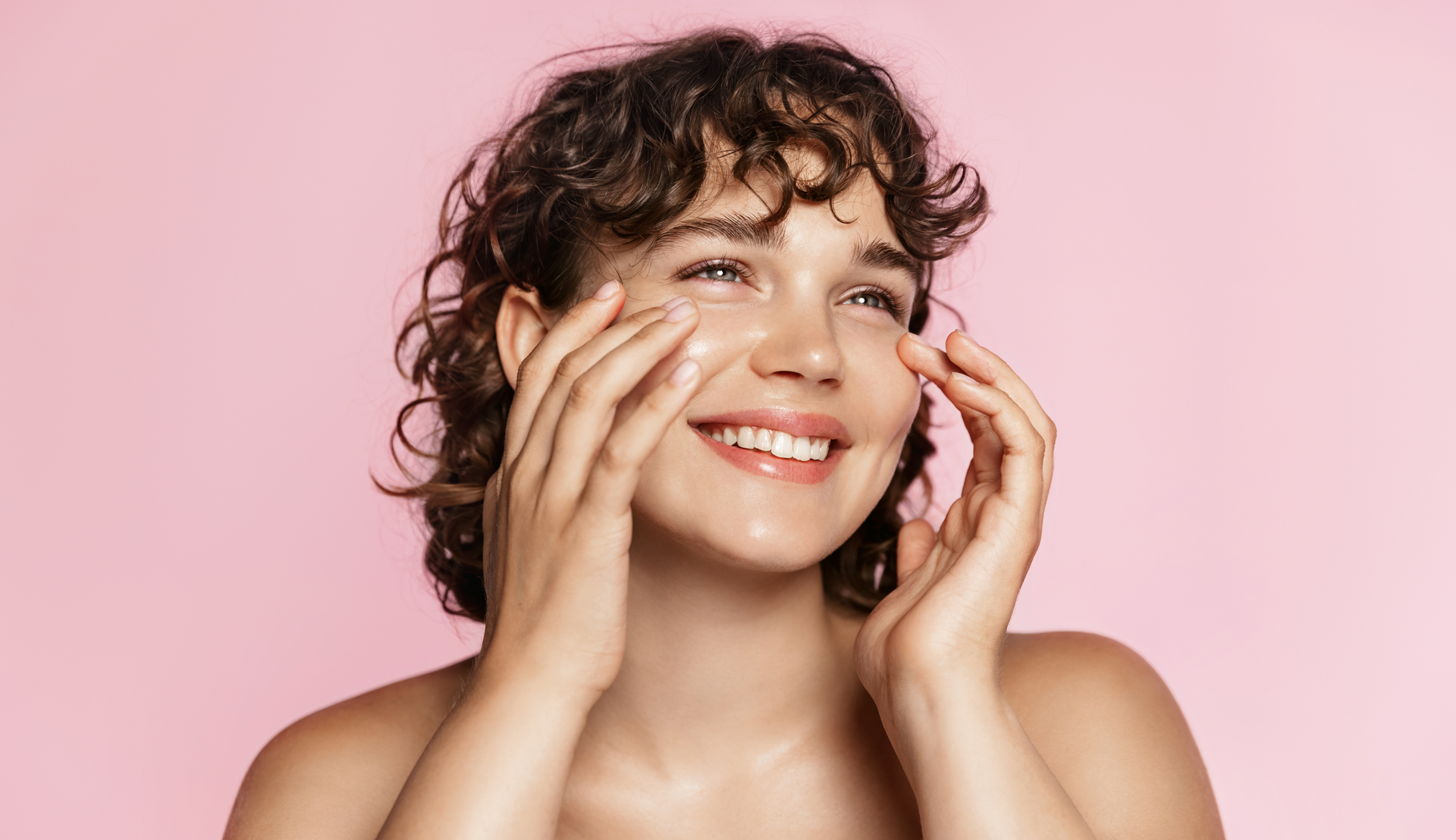 Femme aux cheveux bouclés souriante, appliquant des soins pour le visage avec les deux mains, sur fond rose.
