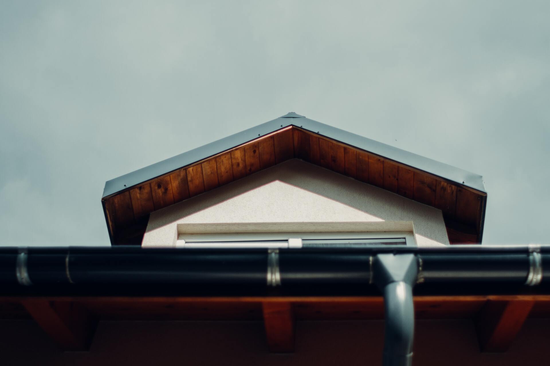 Primer plano del hastial de una casa con un alero de madera y un sistema de canalones oscuros contra un cielo nublado.