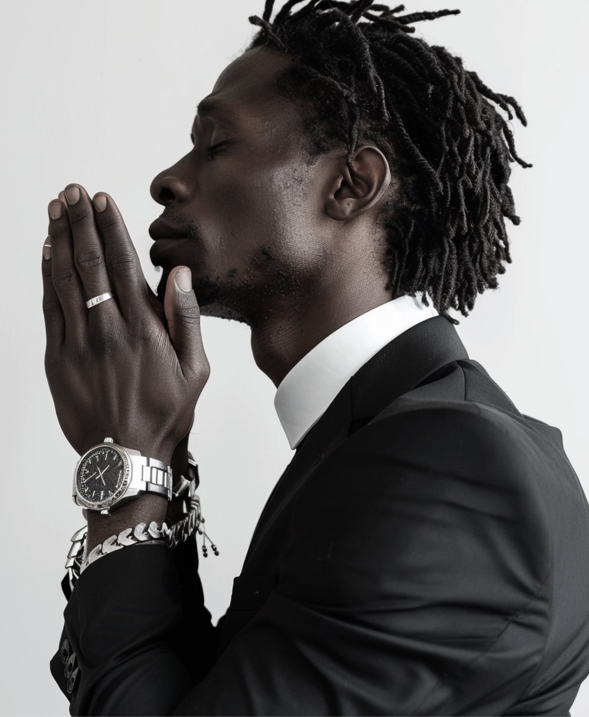 Man in a black suit with hands clasped in prayer, wearing a watch and chain against a light background