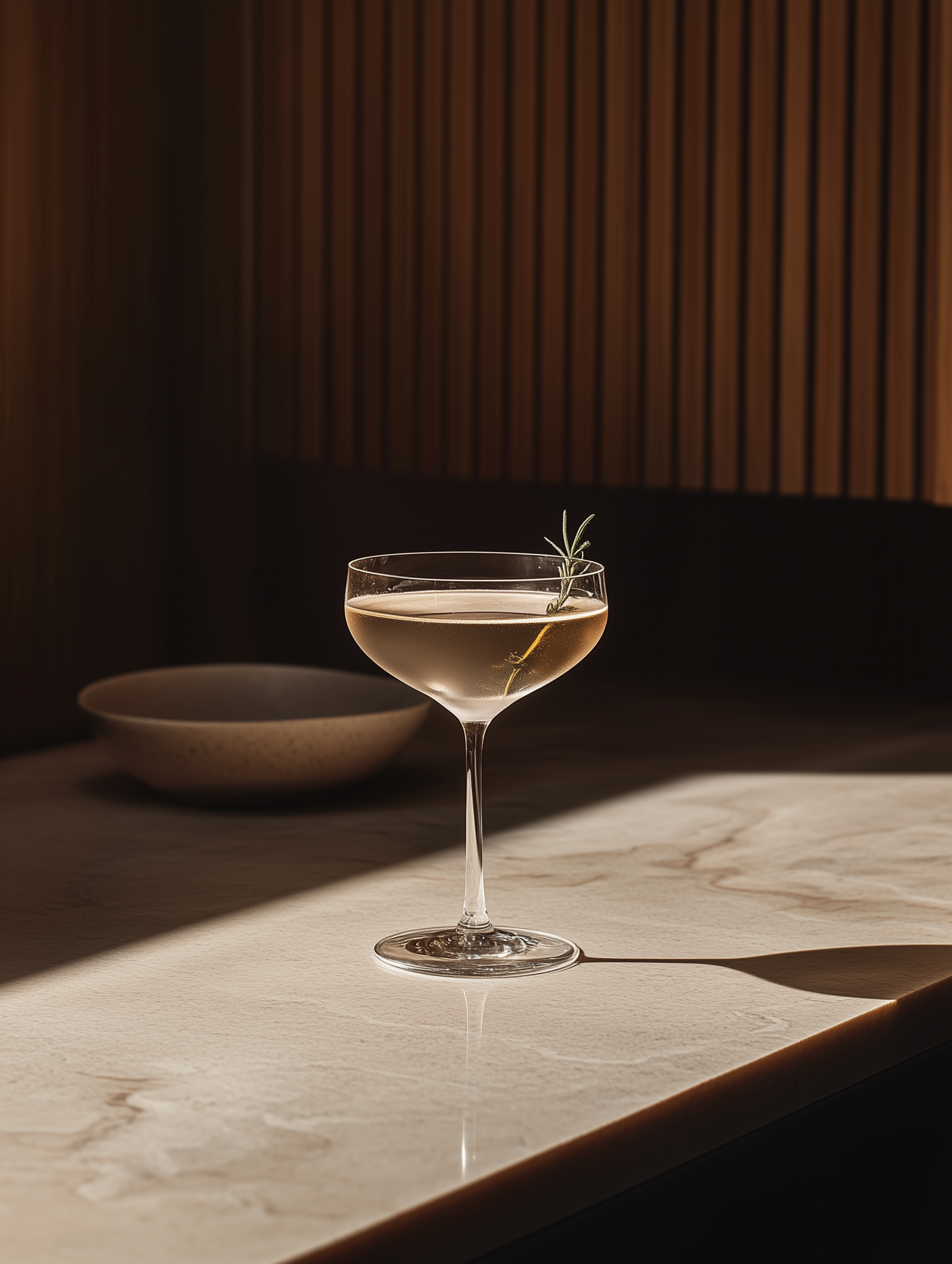 Cocktail in a coupe glass with a sprig of rosemary on a marble countertop, lit by sunlight.