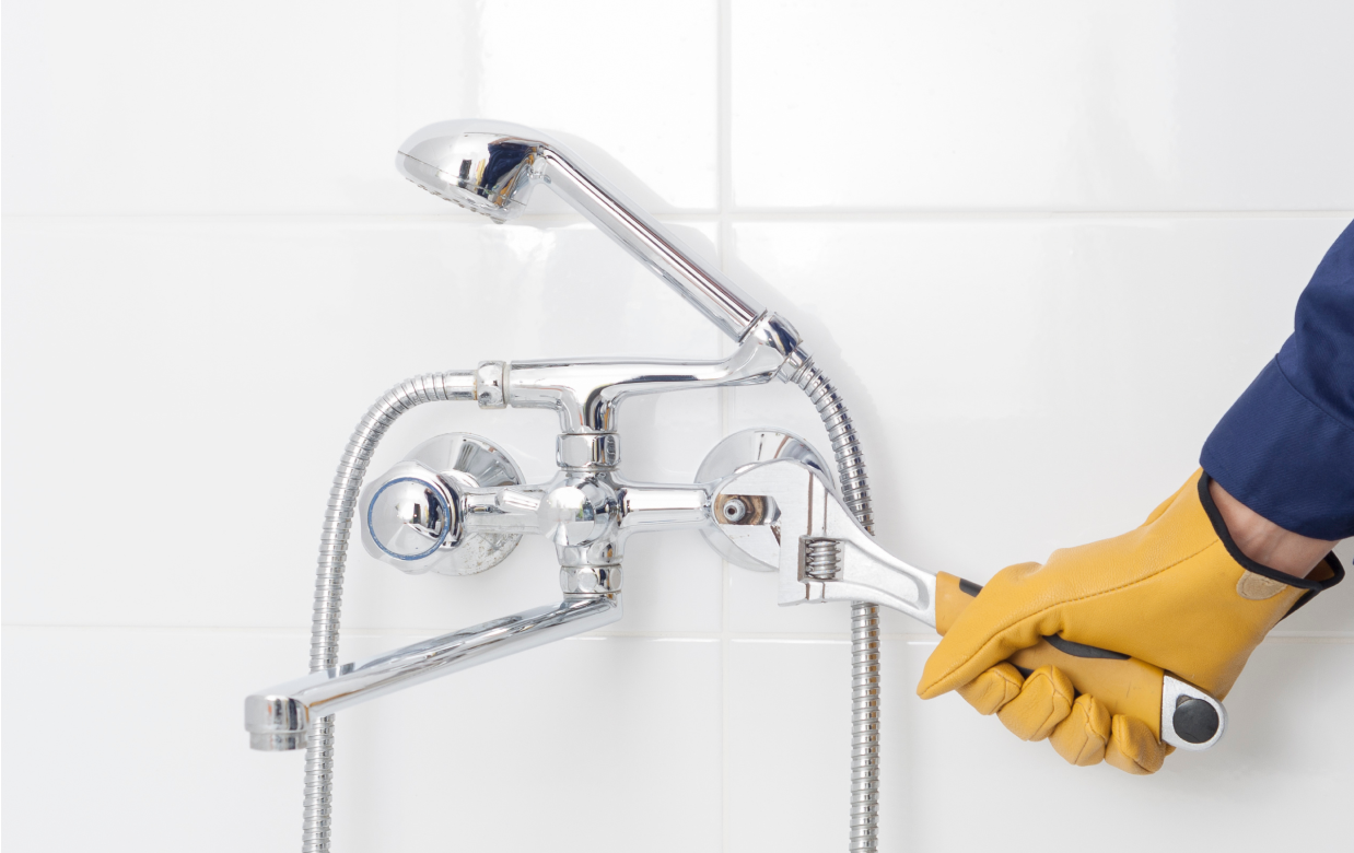 Person in yellow gloves uses a wrench to repair a chrome shower fixture against white tile.