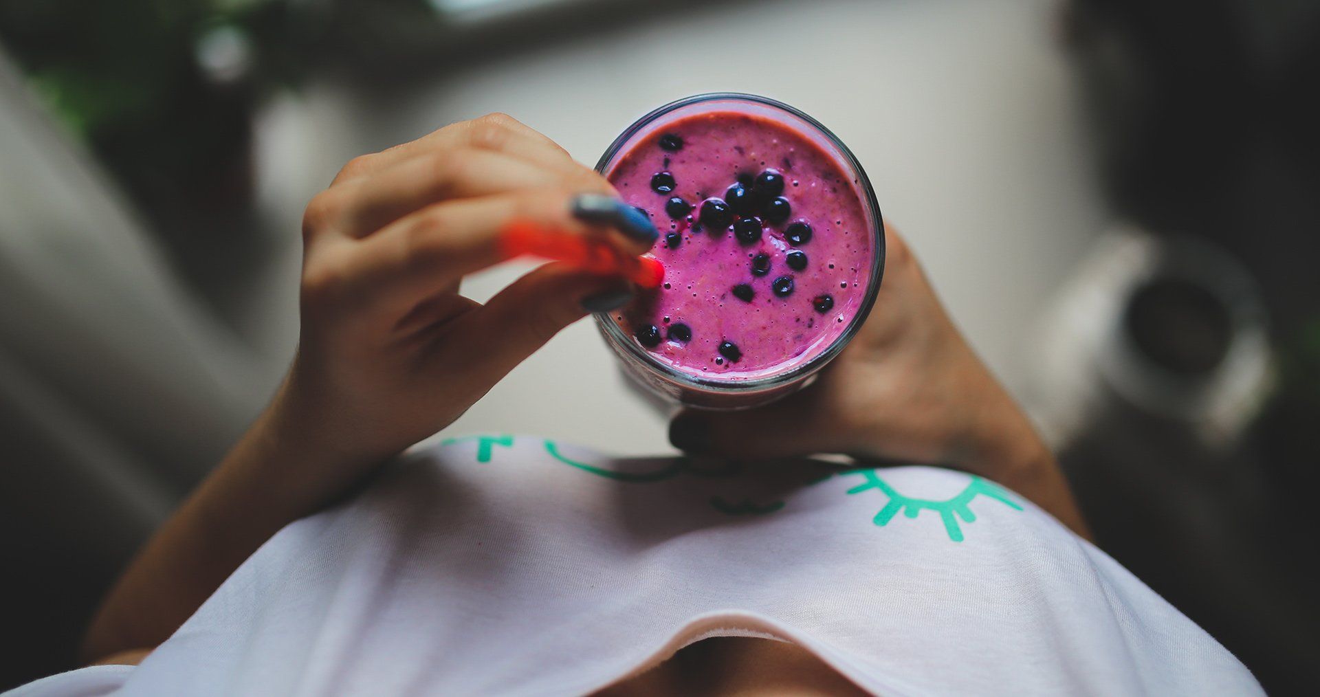 A person holds a glass of bright pink smoothie topped with blueberries, using a straw from a high-angle perspective.