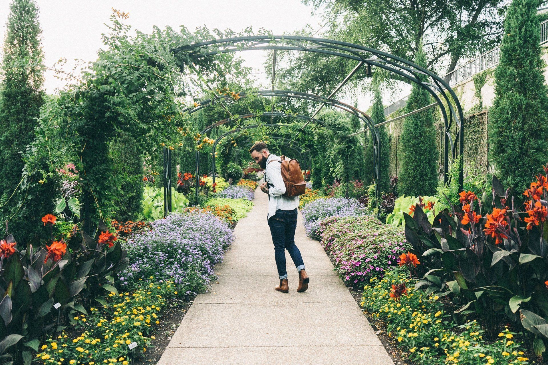 Man with backpack walks down a garden path lined with flowers, under arched trellis.