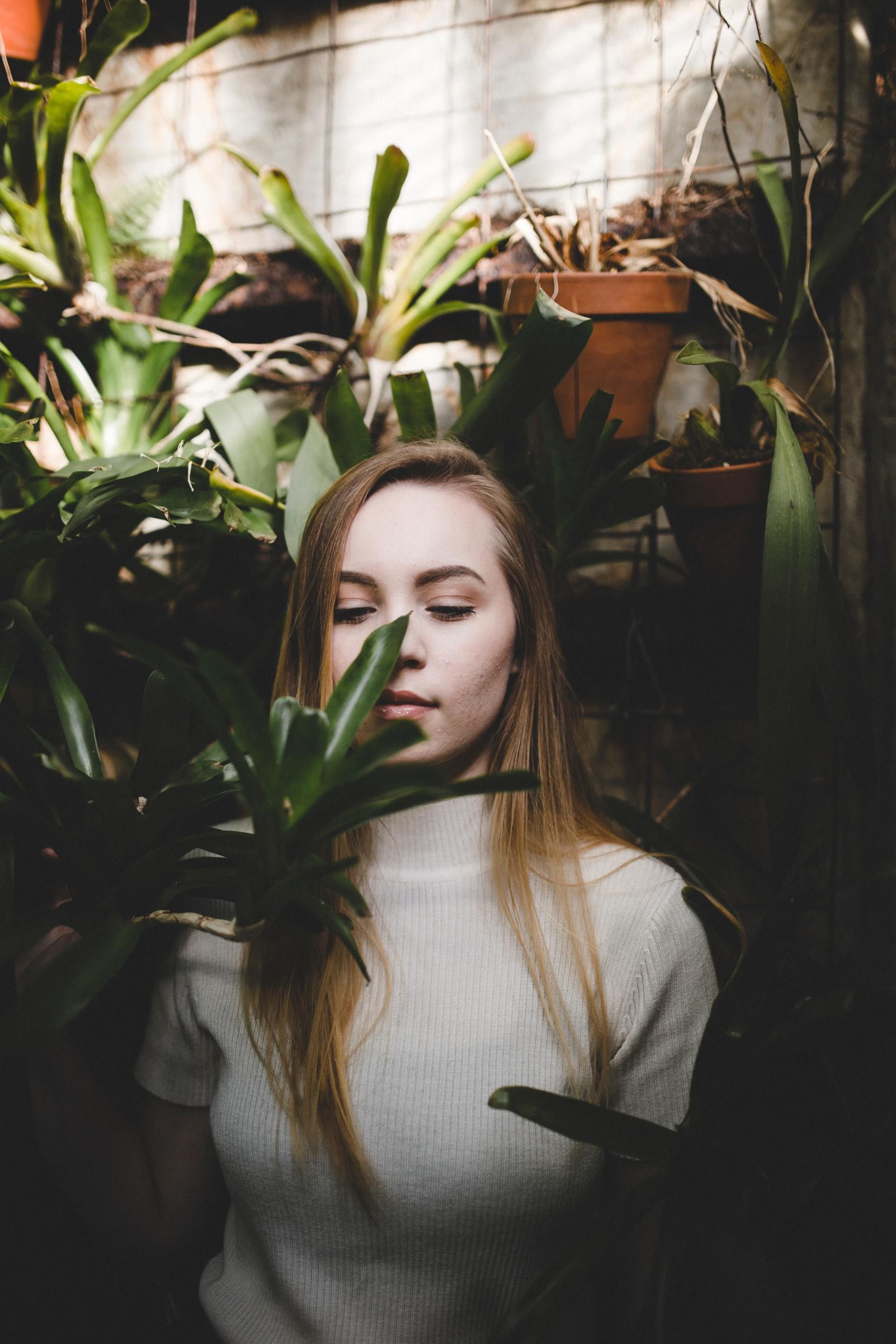 Woman with eyes closed, in a greenhouse, surrounded by green plants, wearing a white sweater.