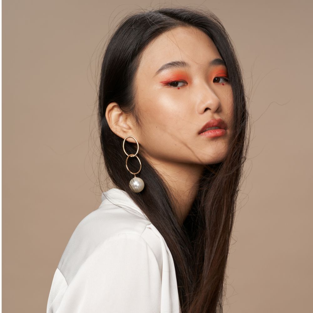 Portrait of a person in a white shirt with orange eye makeup and dangling earrings against a beige background