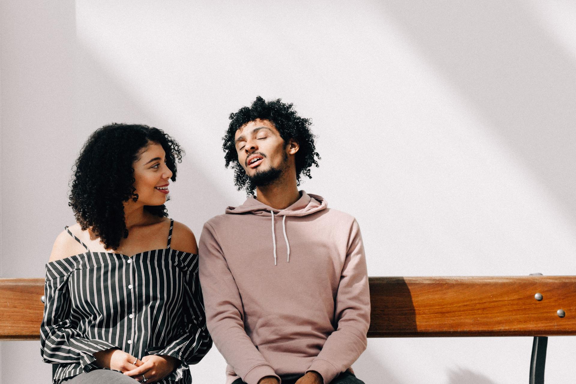 Woman in striped top looks at man in pink hoodie, both seated on a bench against a white wall.