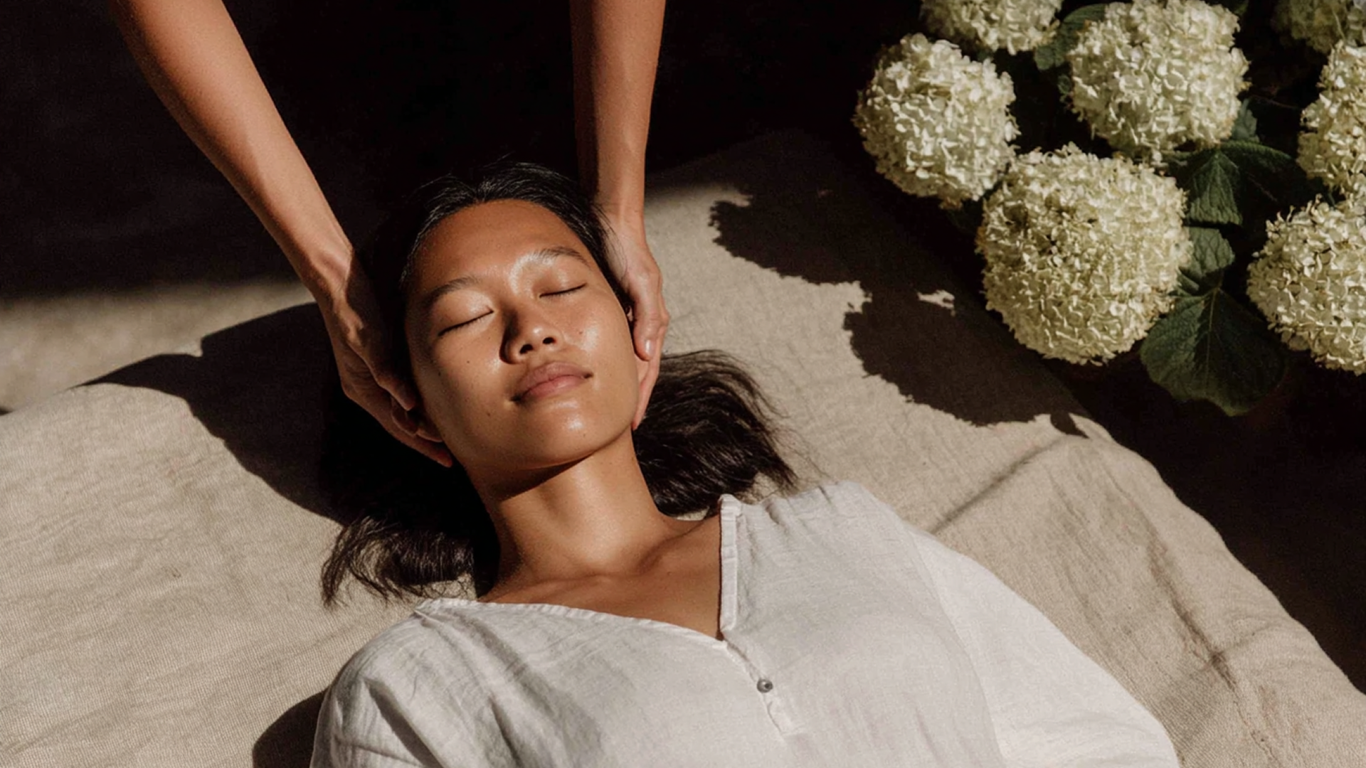 Person reclining on a sunlit bed with eyes closed while another hand massages their head, with white flowers nearby.