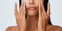 Woman applying skincare product to her face, using hands to massage, neutral background.