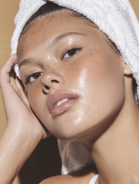 Woman with damp, radiant skin wrapped in a white towel, freckles, looking at camera, gold background.