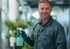 Smiling worker in gray coveralls holding a green spray bottle in a blurred office hallway