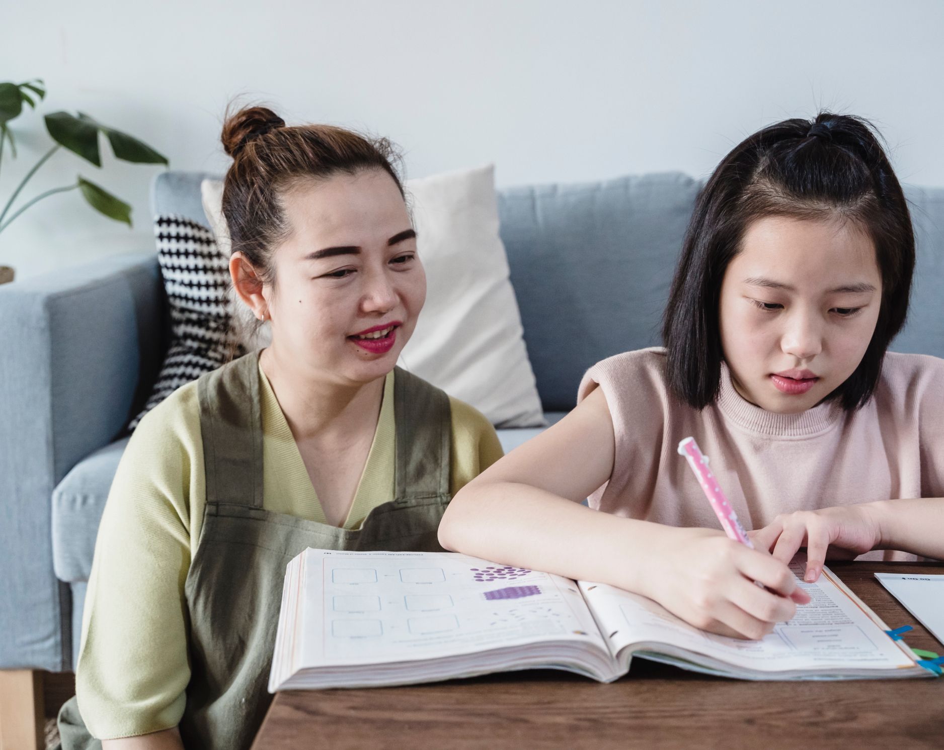 Woman watches a child write in a book at a table; couch in background.