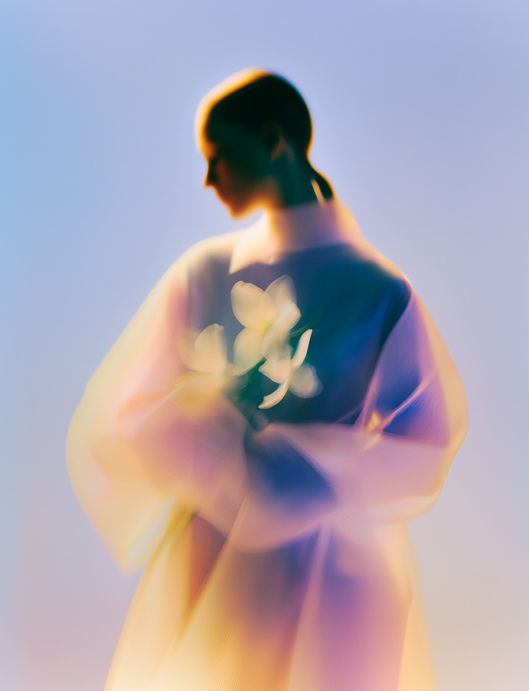 Blurred figure in a flowing pastel garment, viewed from behind against a soft blue-purple background.