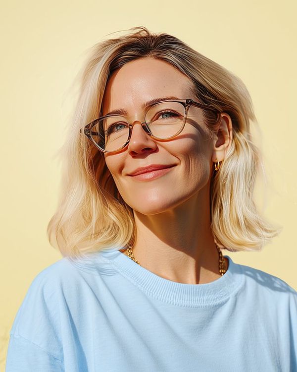 A person with blonde hair and glasses smiles gently against a solid yellow background, wearing a light blue shirt.