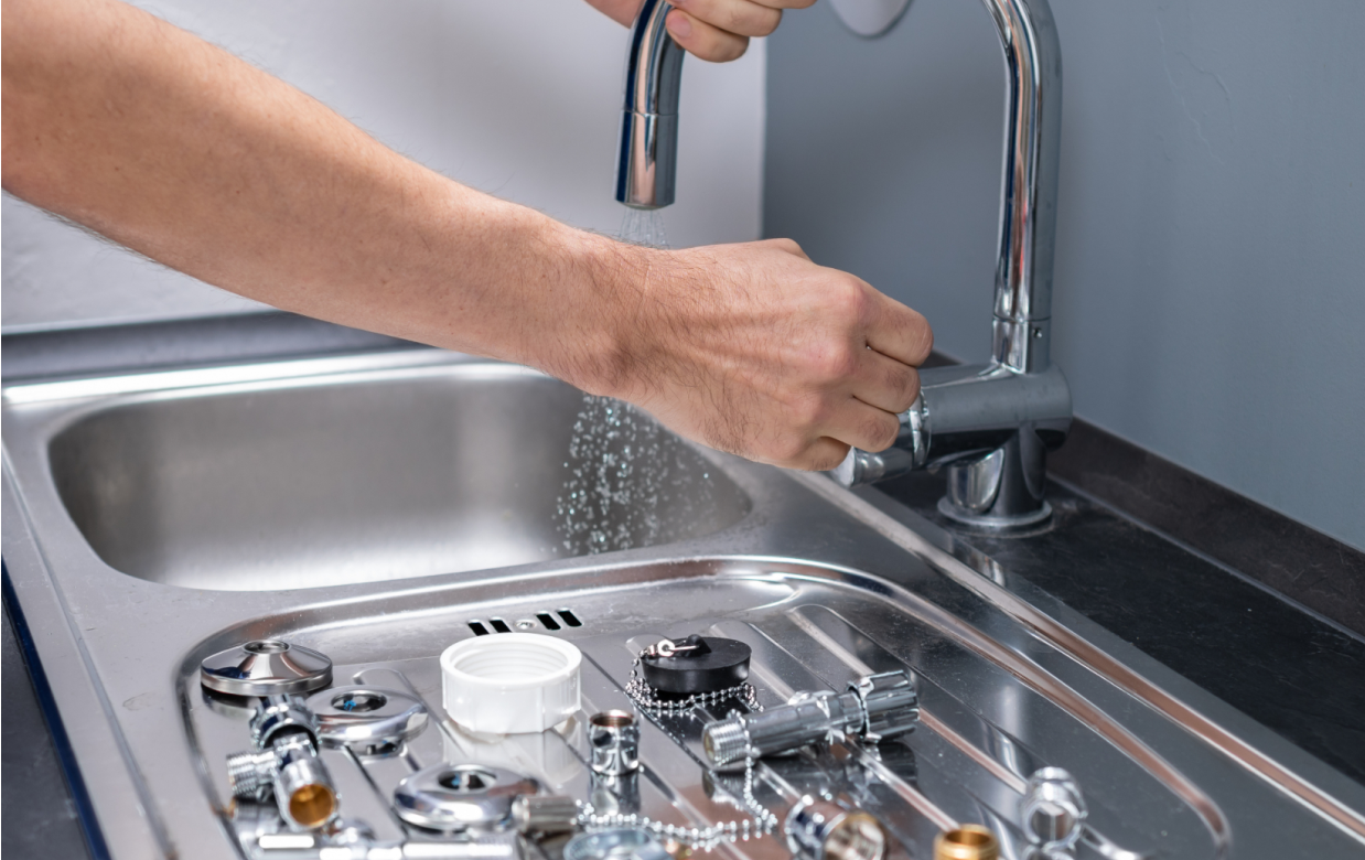 A hand operates a chrome kitchen faucet above a sink containing scattered metal plumbing components.