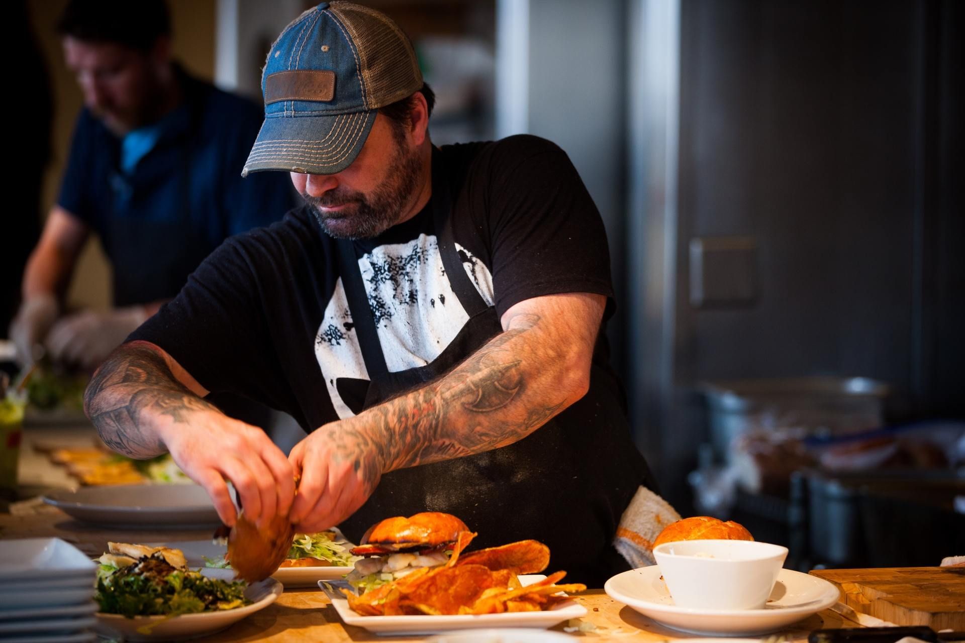 A chef with tattooed arms wears a baseball cap and apron while assembling burgers in a busy kitchen.
