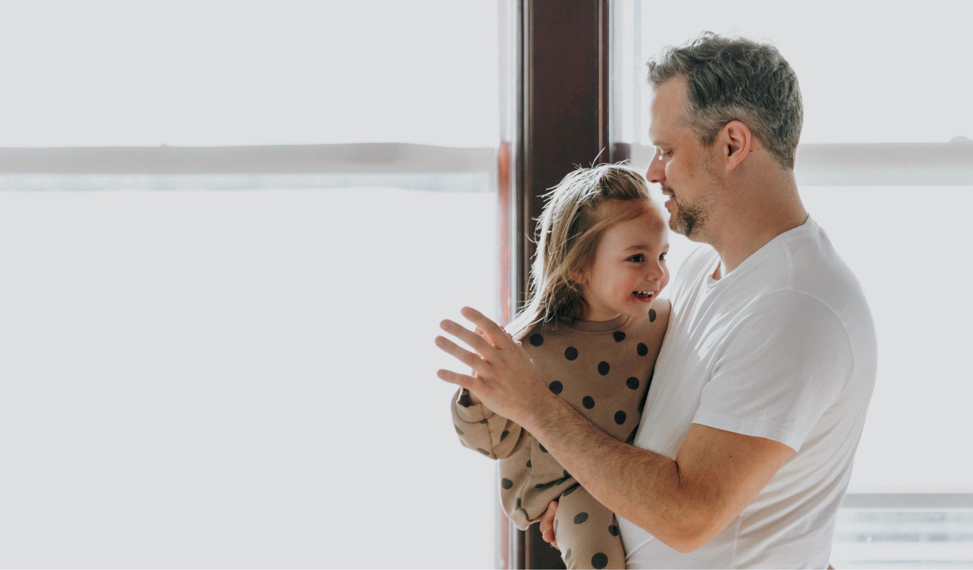 Adult holding a smiling child indoors by a bright window