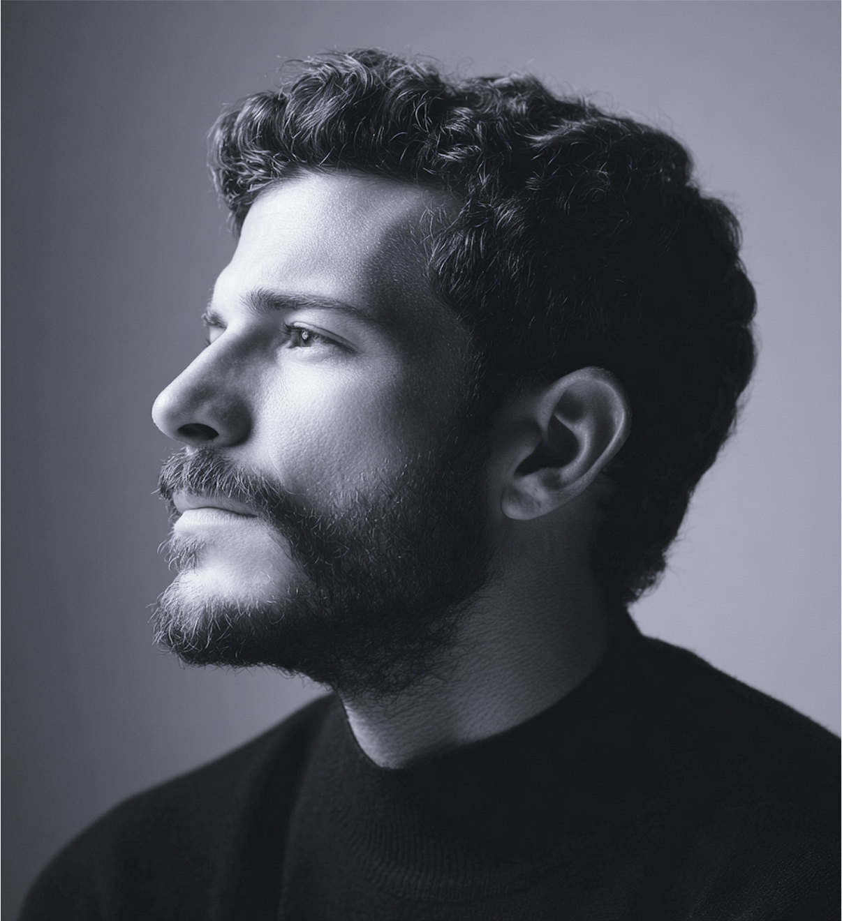 Man in profile, looking upward with a thoughtful expression. Black and white, wearing a dark turtleneck, with beard and curly hair.