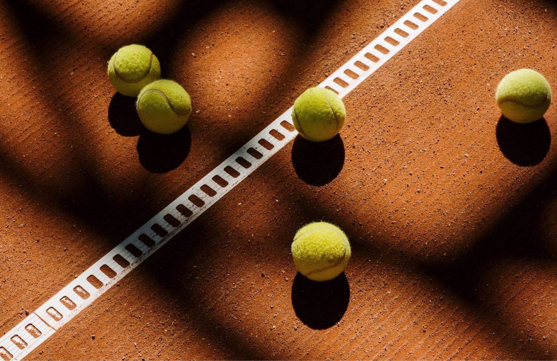 Tennis balls on a red clay court, near a white line, casting shadows.