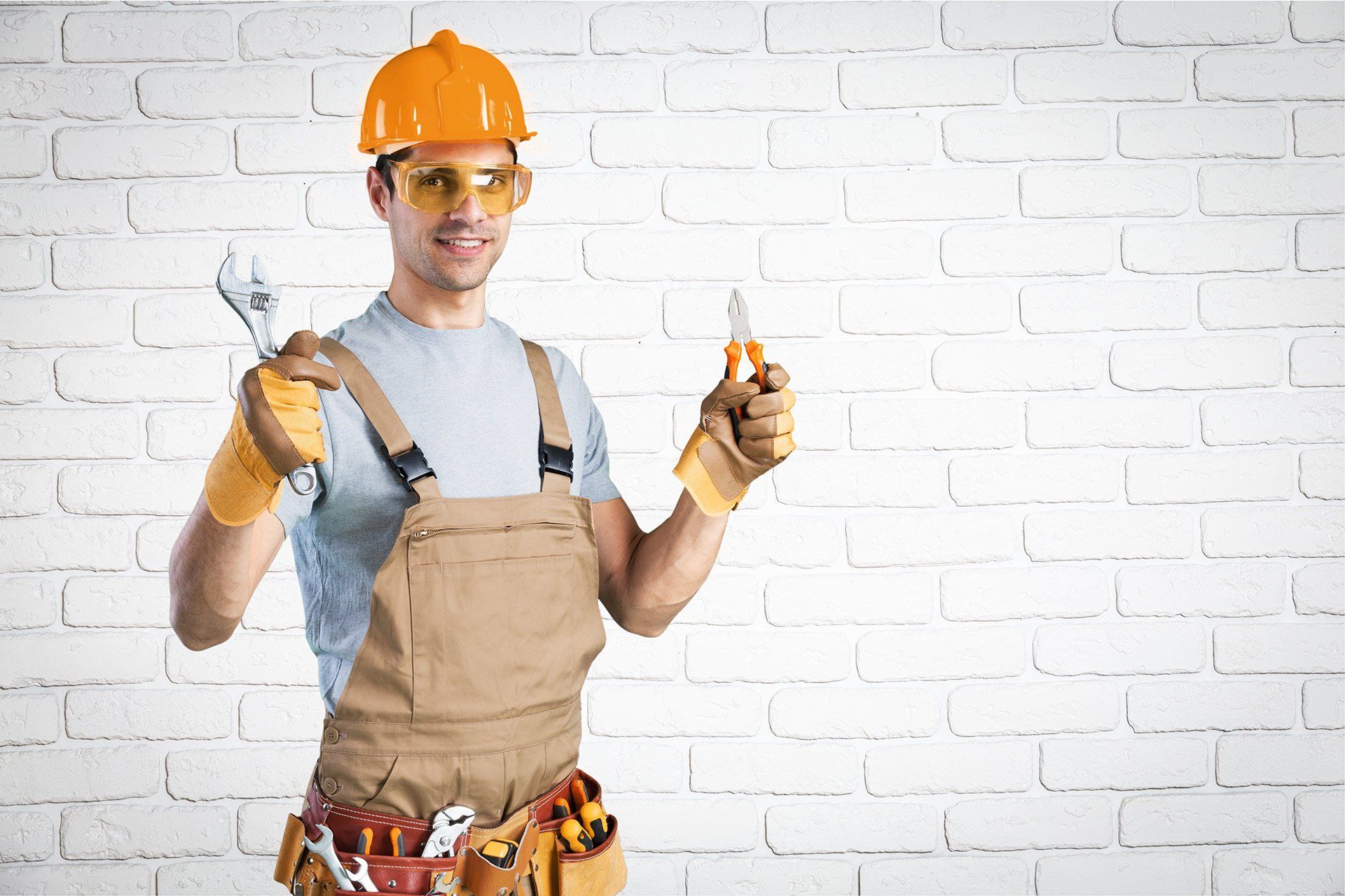 A construction worker is holding a wrench and a screwdriver in front of a white brick wall.