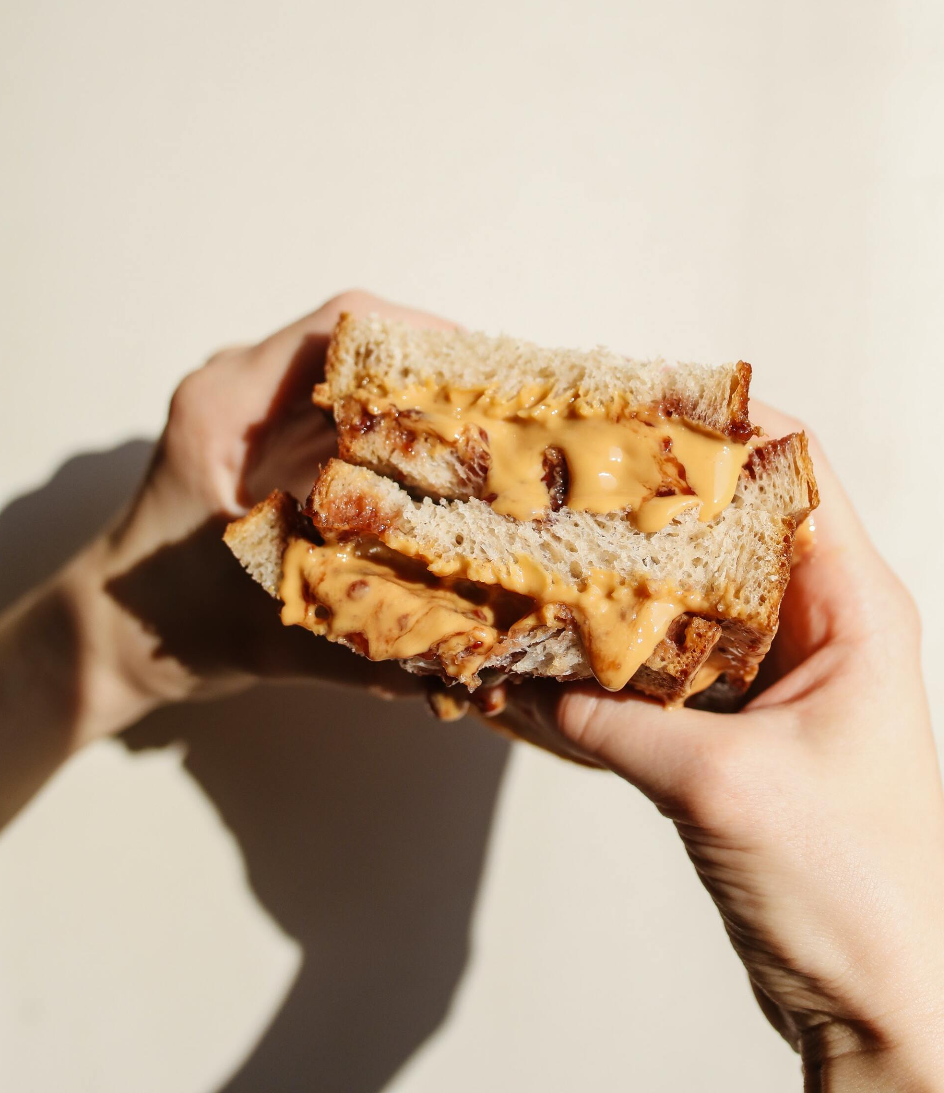 Handen die een doorgesneden pindakaassandwich met beige vulling tussen geroosterd brood vasthouden.
