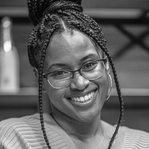 A person wearing glasses and braided hair styled in a bun smiles at the camera, wearing a ribbed top.