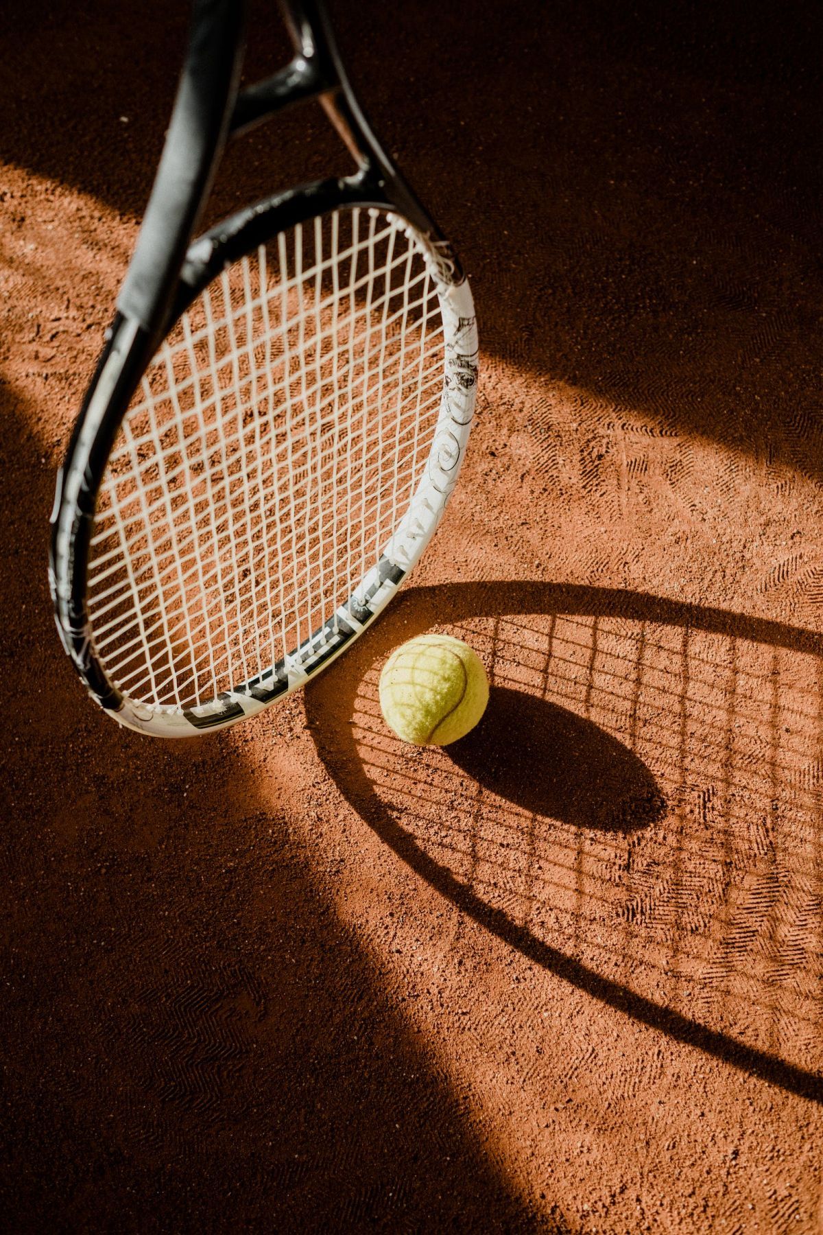 Tennis racket and ball casting shadows on a clay court under warm sunlight