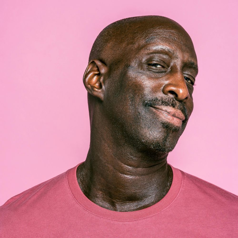 Man in a pink shirt posing against a pink background, head tilted with a slight smile