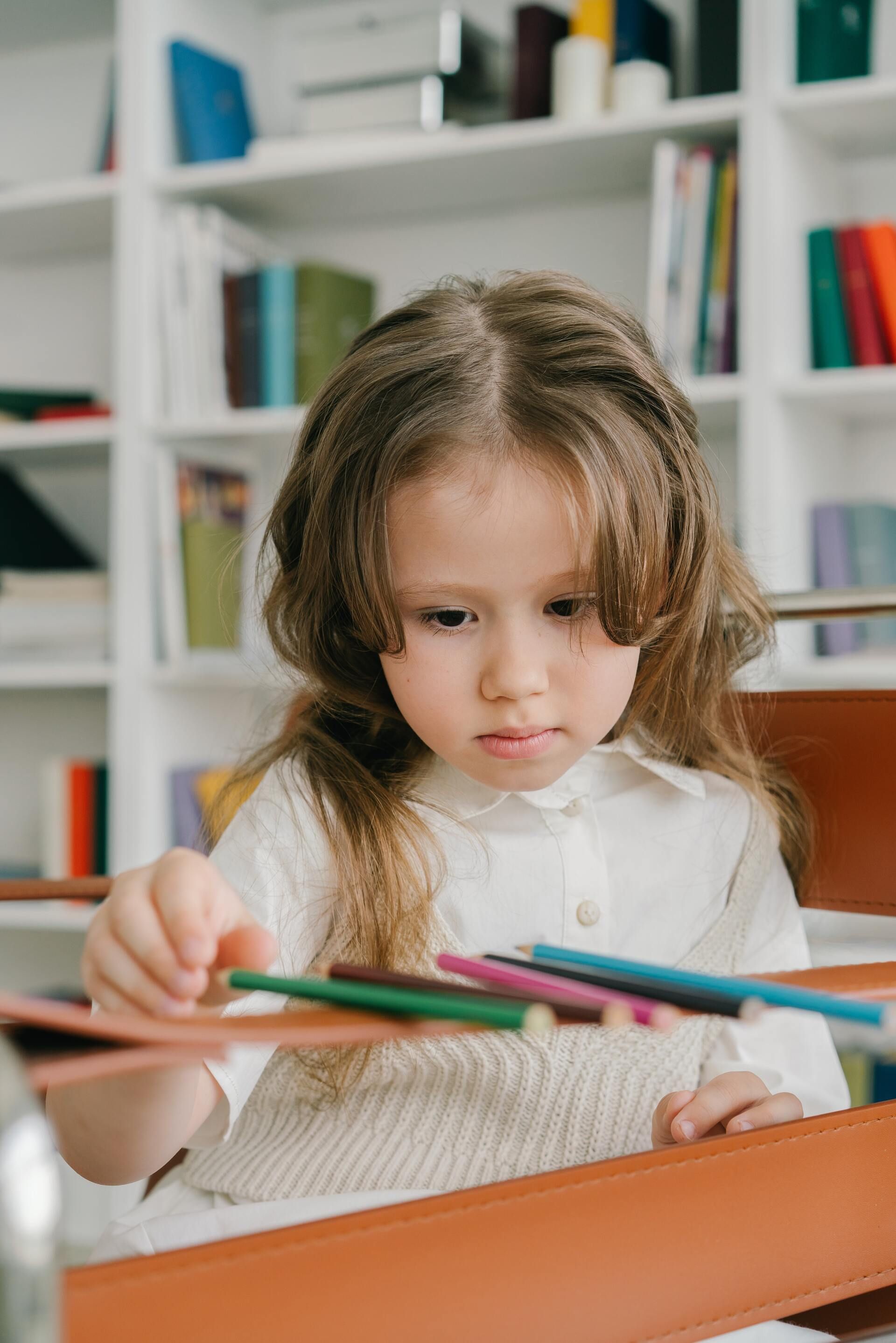 Child drawing with colored pencils at a table, concentrating in a bright room with shelves in the background