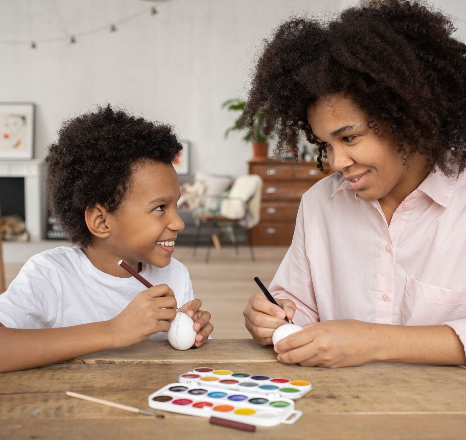 Two people painting eggs at a table with colorful watercolor paints, smiling in a bright room