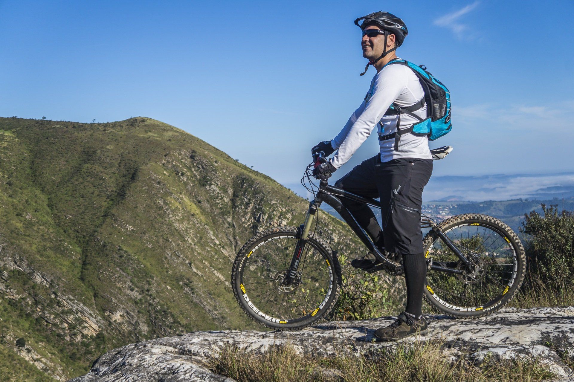 Man on mountain bike, looking away from camera, standing on a mountaintop, sunny sky, with mountain view.