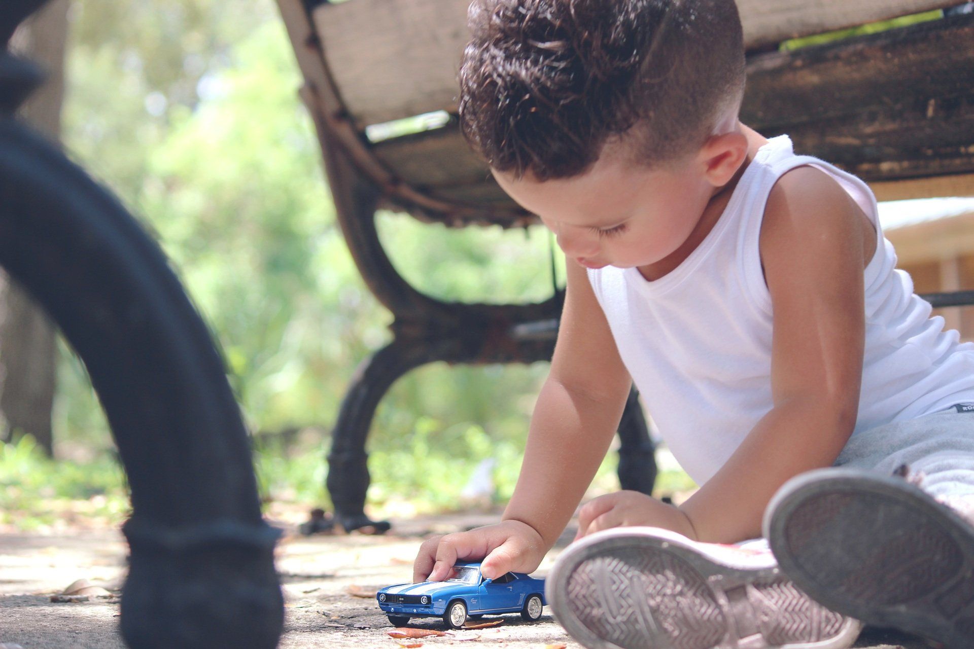 Child playing with a blue toy car on a concrete surface near a bench outdoors.