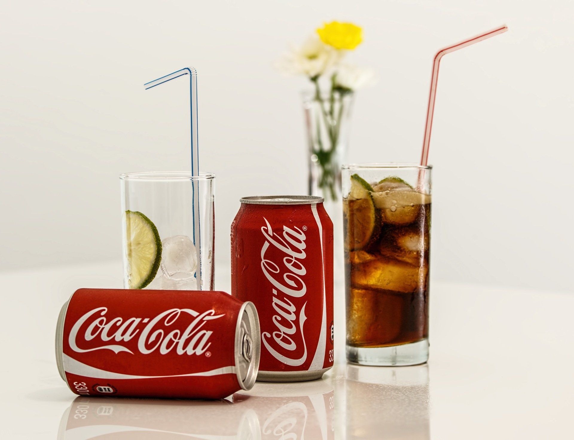 Two Coca-Cola cans, a glass of cola with lime and ice, and an empty glass with lime and a straw on a table.
