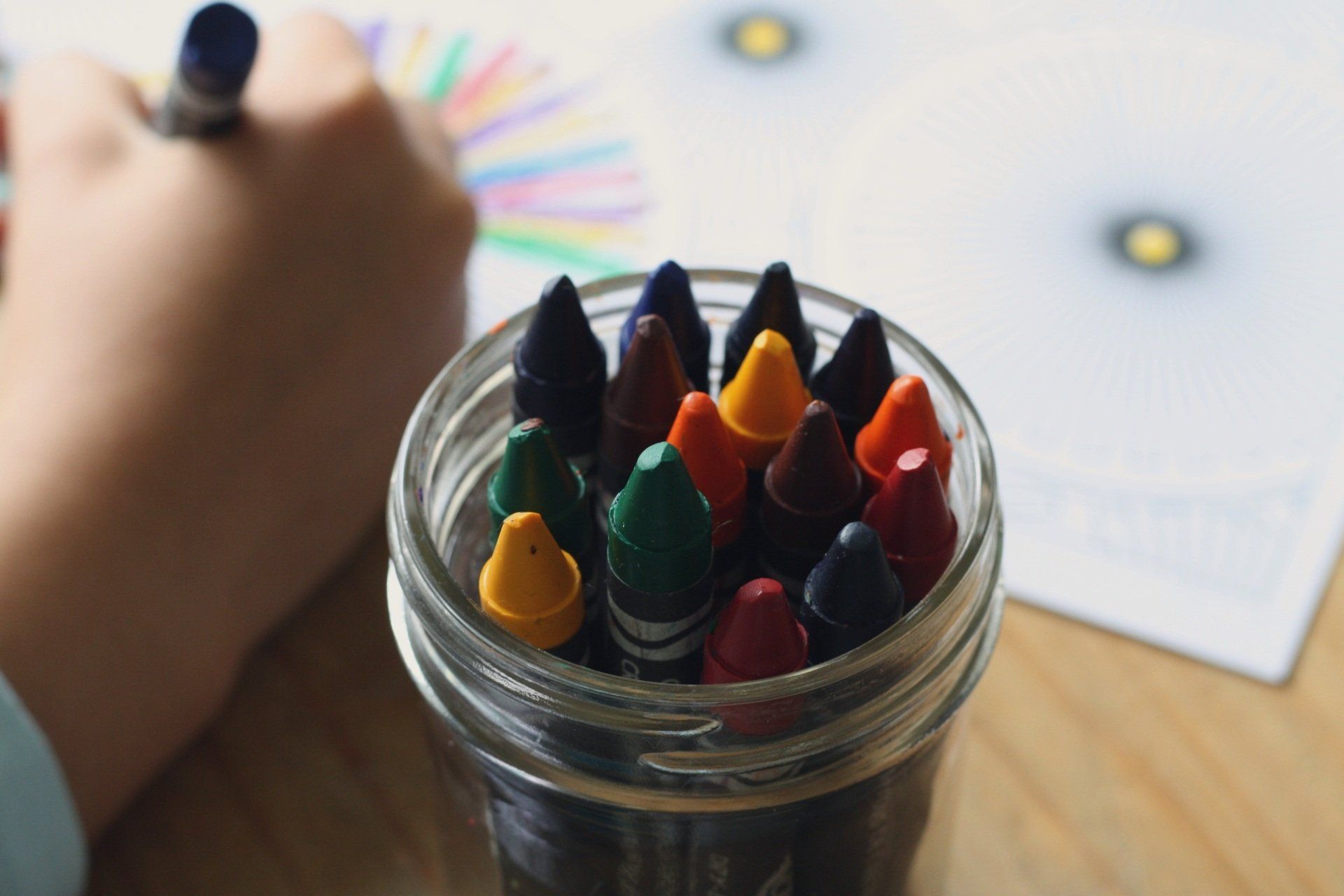 A mason jar full of sharpened crayons in focus, child hand is drawing on paper in background and slightly out of focus