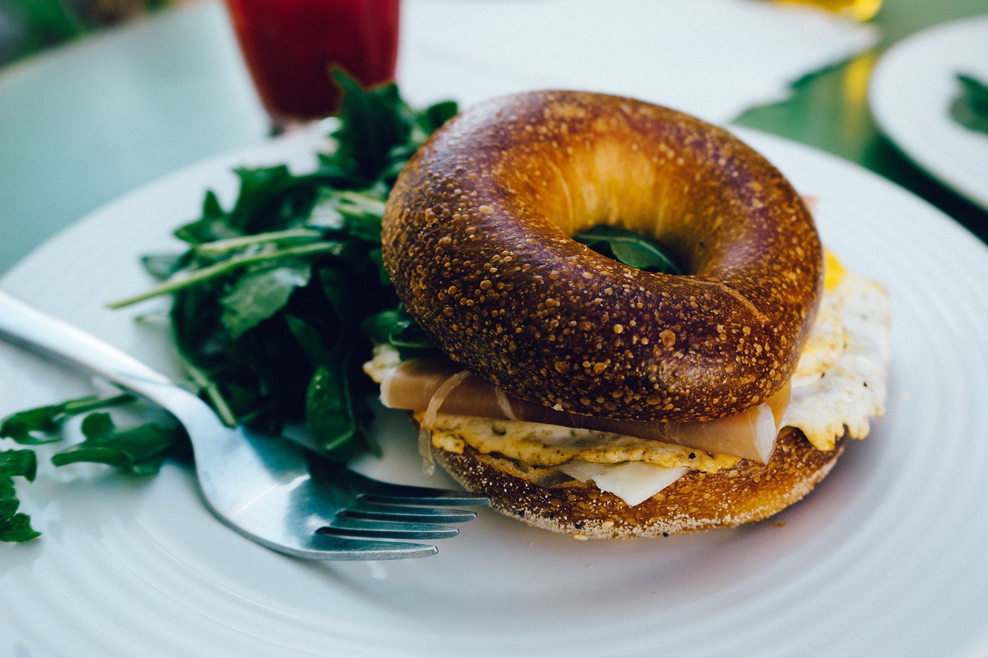 Bagel sandwich with egg and ham on a plate with salad and fork.