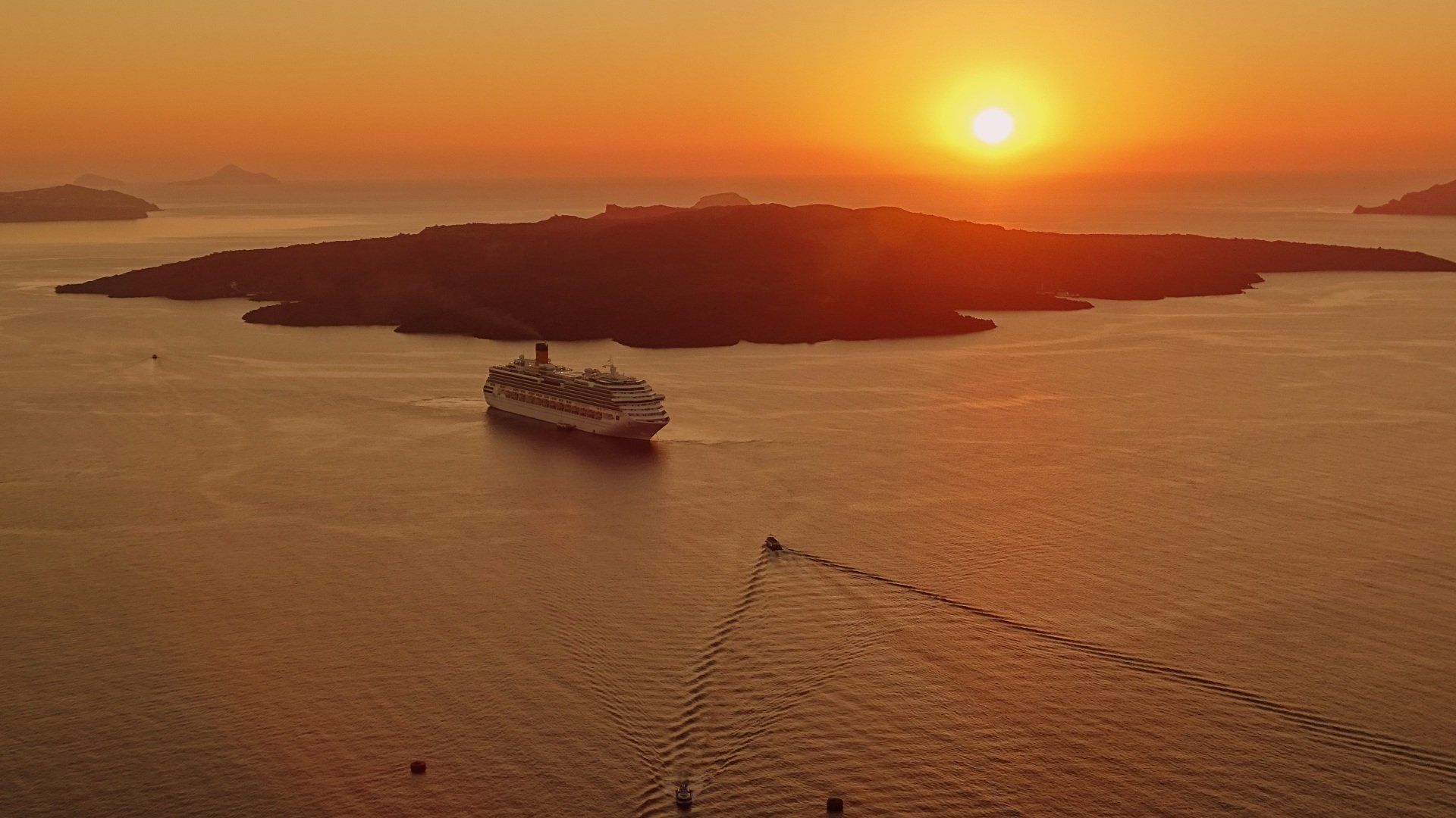 Sunset over Greek island, cruise ship sails on calm water, warm orange hues.
