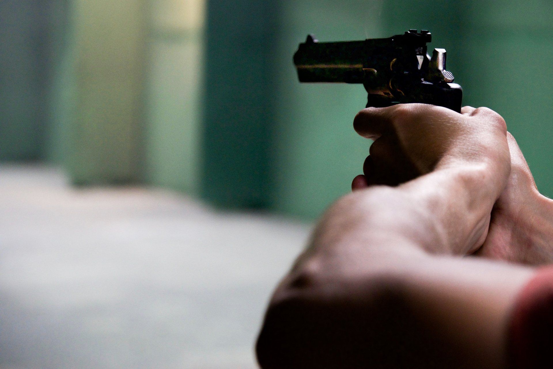 A person’s arms are extended, holding a handgun in a two-handed grip, aimed forward in a green-toned indoor setting.