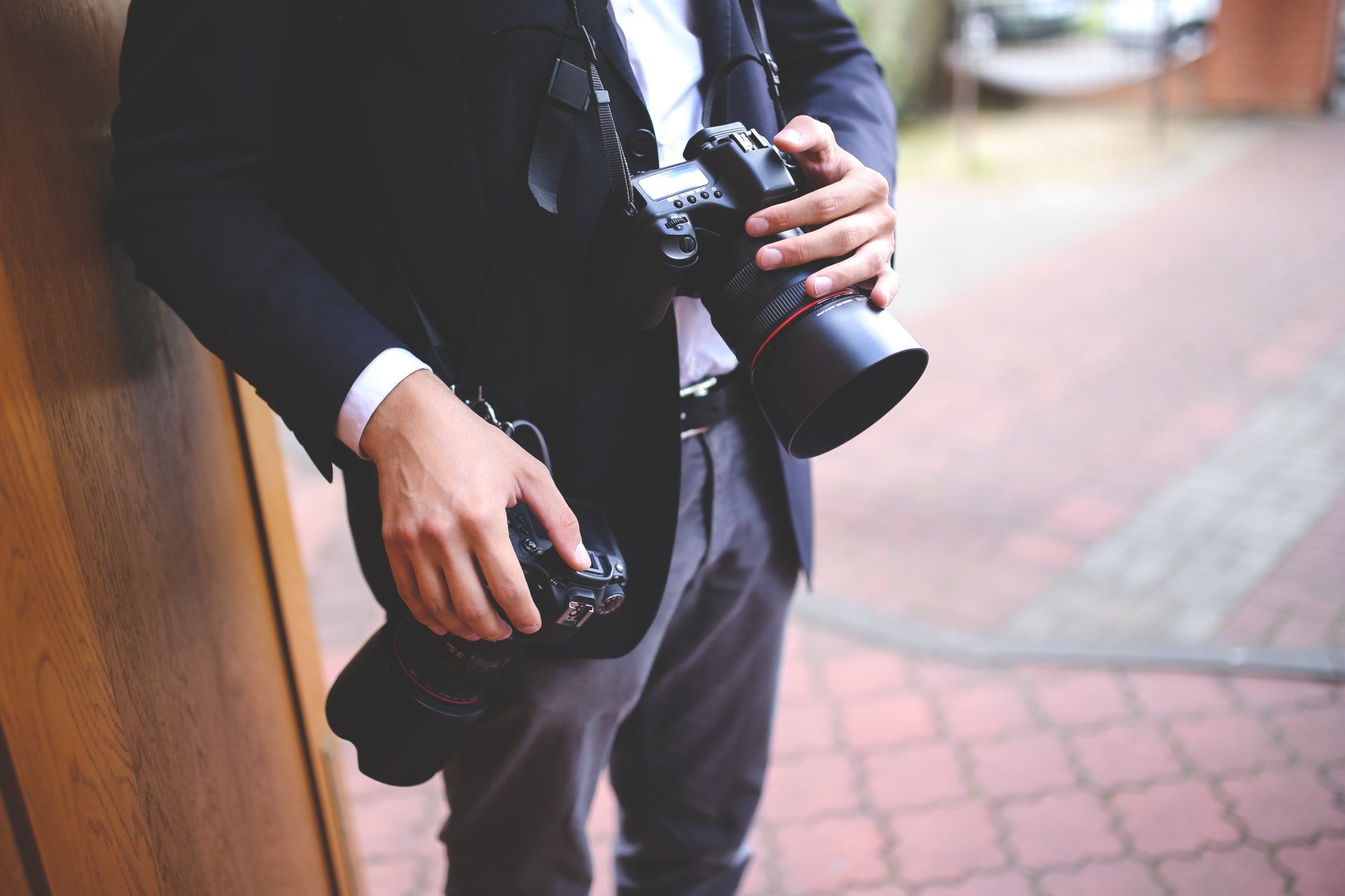 Person in a black suit holding a camera beside a brick walkway